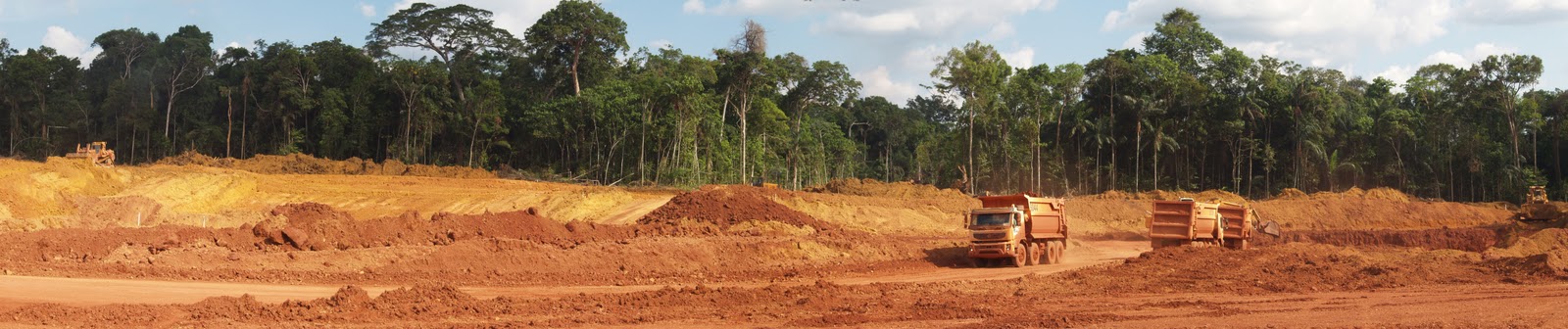 Raw earth has been stripped of trees leaving a green canopy seen in the horizon revealing in the foreground swathes of reddish light brown earth and on the far right are big trucks moving and hauling dirt.