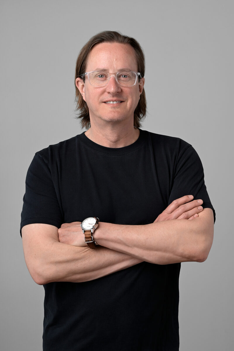 A white man stands in front of a medium gray background, wearing a black tshirt with his arms crossed. He has clear-frame glasses on and a wristwatch with a brown band.