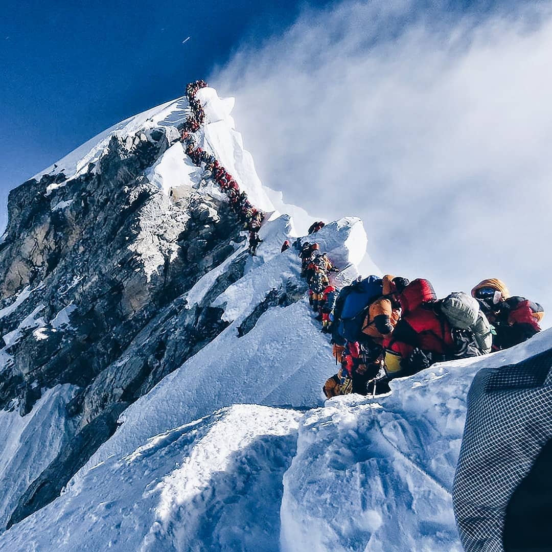 Close up of peak of snowcapped mountain, with a line of people in colorful jackets climbing to the peak.