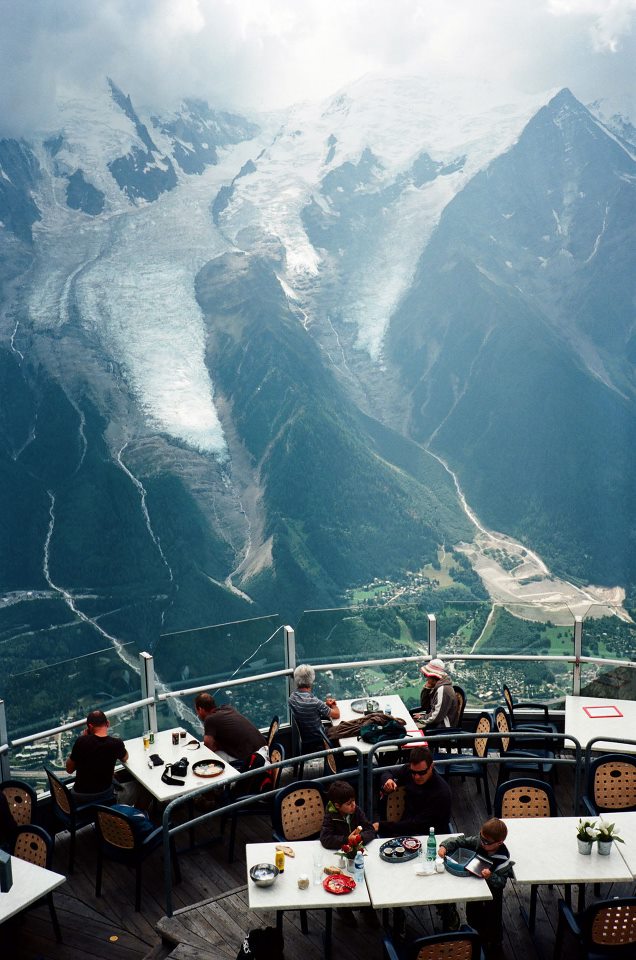 Rectangular landscape with giant blue-green mountains with snowcaps in the background and the forefront including people dining at a restaurant.
