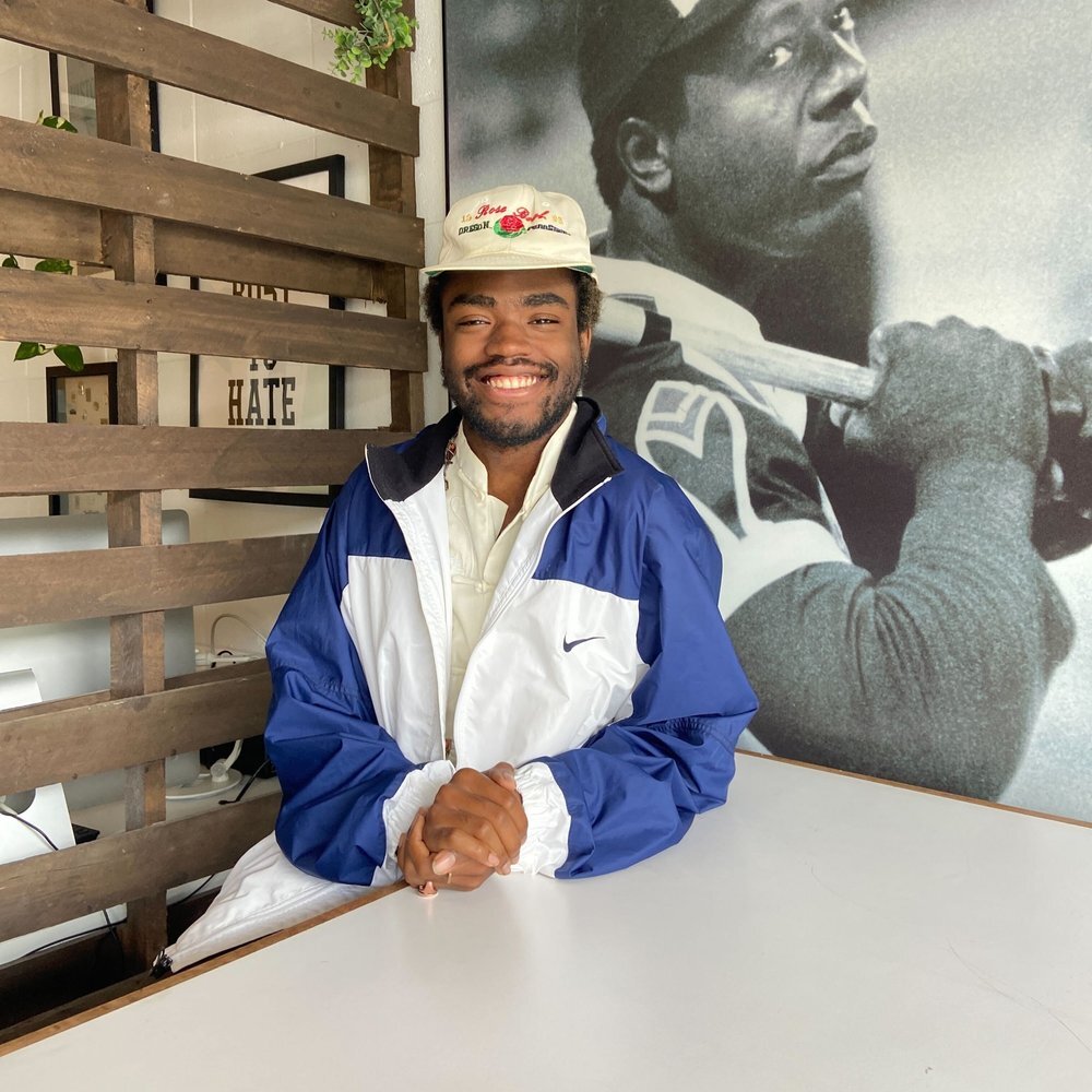 portrait of a person wearing a hat and blue and white jacket, smiling at the camera while holding their hands
